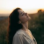 A serene woman with head tilted back, savoring the warm sunset in a field.