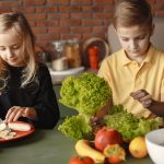 Focused little girl and boy sitting at desk with assorted vegetables and fruits while cooking in loft kitchen with brick wall